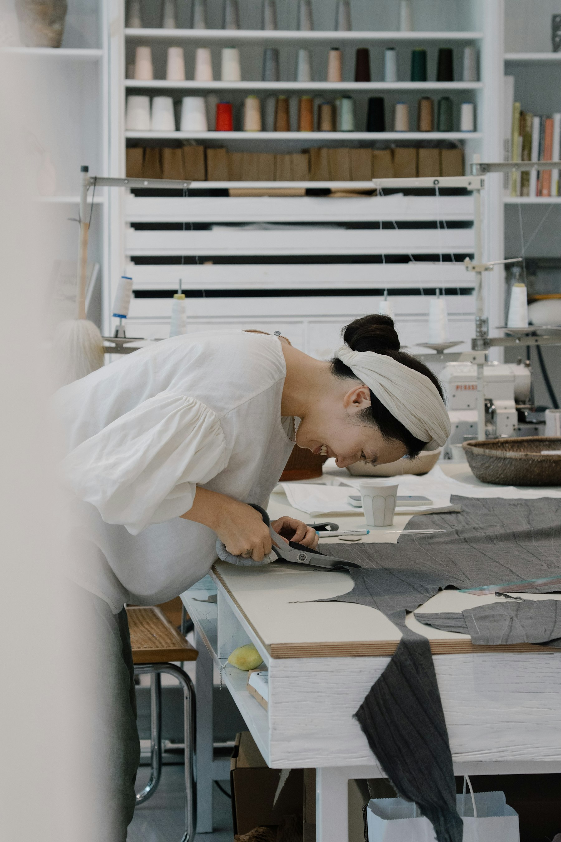 Una mujer trabaja una tela gris en la mesa de corte de un taller, con conos de hilo en las estanterías — imagen ilustrativa